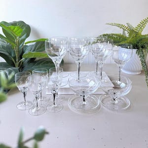 May include: A selection of clear crystal glassware, including wine glasses, champagne coupes, and smaller stemmed glasses. The glasses feature detailed stem designs and are arranged on a white surface, with plants in the background.