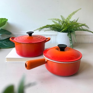 Peut inclure: Deux récipients de cuisson en fonte émaillée avec un dégradé orange vif. L'un est une cocotte ronde avec deux poignées latérales et un couvercle à bouton noir. L'autre est une casserole avec un long manche et un couvercle assorti à bouton noir.