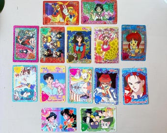 Puede incluir: Una colección de cartas coleccionables de Sailor Moon. Cada carta presenta ilustraciones coloridas de personajes de la serie de anime. Las cartas tienen un acabado brillante y están dispuestas sobre una superficie clara.