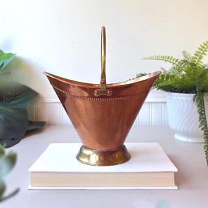 Peut inclure: Un seau à charbon en cuivre poli avec une base en laiton et une poignée verticale unique en laiton. Le récipient présente une large ouverture évasée et un détail de bordure perlée décorative le long du rebord supérieur en métal.