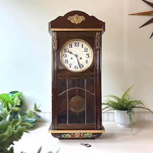 Puede incluir: Un reloj de pared de madera marrón oscuro con una esfera blanca circular y números arábigos negros, tallas decorativas doradas en la parte superior y los lados, una sección inferior acristalada con un péndulo de latón y detalles florales pintados.