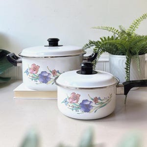 Può includere: Due pentole in smalto bianco con coperchi e manici neri, caratterizzate da un motivo floreale con fiori viola, rosa e blu su steli verdi. La pentola più grande ha due manici laterali, mentre il pentolino ha un lungo manico nero.