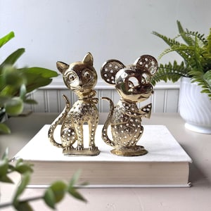 May include: Two gold-colored figurines: a cat and a mouse. The cat is seated with a bow tie, and the mouse holds a cheese wedge. Both are embellished with small, white dots. They are displayed on a white book.