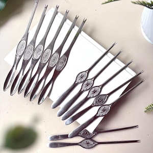 May include: A set of twelve silver-toned metal seafood picks, each featuring a two-pronged tip and a central oval medallion embossed with a detailed crustacean design. The slender handles taper into a smooth, rounded spoon-like end for serving.