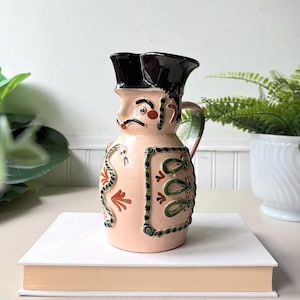 Peut inclure: Un pichet en céramique en forme de personnage portant un tricorne noir. Le pichet présente une base couleur pêche clair avec des détails peints à la main, notamment un serpent vert, des motifs floraux rouges et un panneau rectangulaire.
