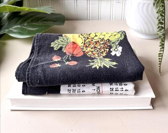 May include: A folded black towel with a colorful pineapple and floral design, resting on a white book. A small calendar with the month of November is visible. The towel features red, yellow, and green accents.