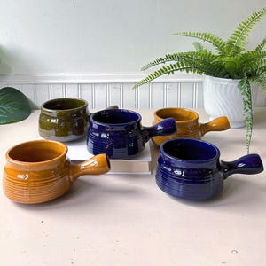 May include: A collection of five ceramic soup bowls with handles, featuring a ribbed texture on the lower exterior. The bowls are finished in glossy glazes of mustard yellow, deep navy blue, and olive green, arranged in a cluster on a flat surface.