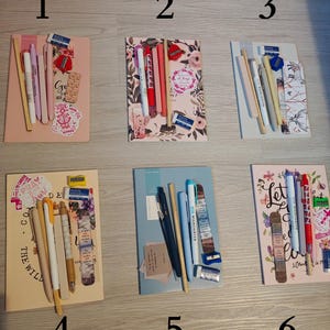 Op de afbeelding: Zes notitieboekjes met briefpapier zijn gerangschikt. Elk notitieboekje heeft pennen, potloden, markeerstiften, gummen en stickers. De notitieboekjes zijn in roze, blauw en beige, met bloemen- en inspirerende ontwerpen.