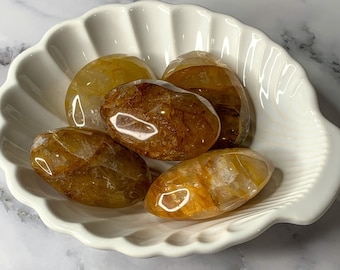 May include: Five tumbled citrine stones in a white ceramic shell-shaped dish. The stones are a light yellow color with brown inclusions.