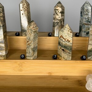 May include: Eight green, brown, and white striped crystal points arranged on a wooden shelf. The points are numbered 1 through 8. Four clear quartz stones are in the bottom right corner of the image.