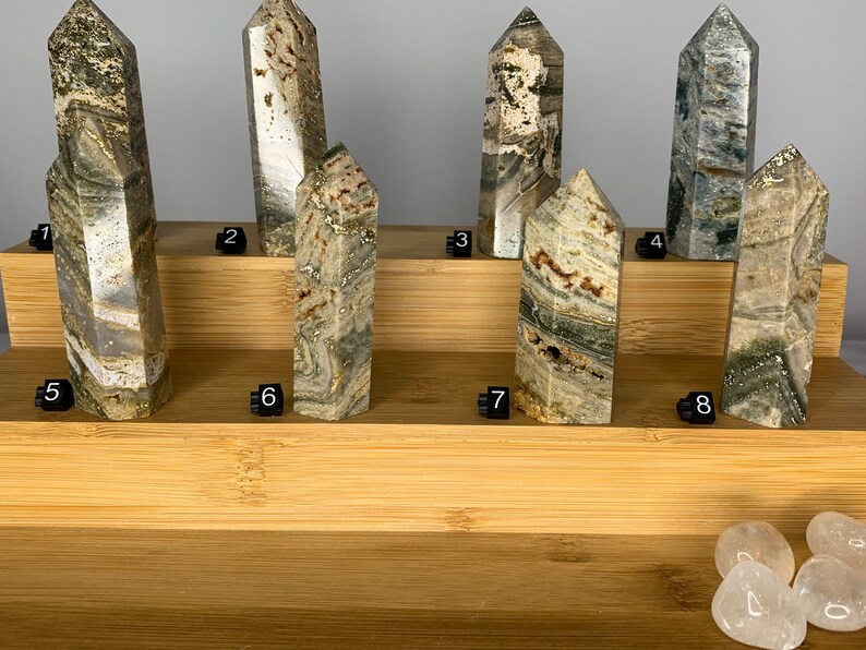 May include: Eight green, brown, and white striped crystal points arranged on a wooden shelf. The points are numbered 1 through 8. Four clear quartz stones are in the bottom right corner of the image.