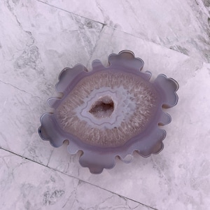 May include: A polished agate geode slice with a scalloped edge. The geode displays concentric bands of light purple, white, and grey hues, with a central cavity lined with sparkling crystals. The natural stone is displayed on a marble surface.