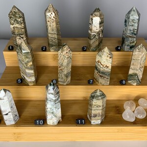 May include: Eleven green, brown, and white striped crystal points arranged on a tiered bamboo display stand. Four clear quartz stones are on the bottom shelf.
