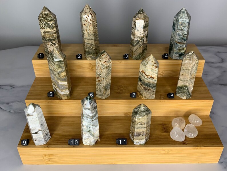 May include: Eleven green, brown, and white striped crystal points arranged on a tiered bamboo display stand. Four clear quartz stones are on the bottom shelf.