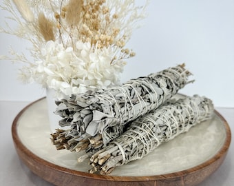 May include: Two bundles of white sage smudge sticks on a wooden tray with a white background. The sage is tied with twine and the bundles are arranged in a criss-cross pattern.