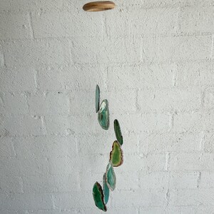 Op de afbeelding: Een hangende mobiel met een houten bovenstuk en een reeks groene en blauwgroene agaat schijfjes. De agaat schijfjes, in verschillende maten en vormen, zijn opgehangen aan een dun touwtje, wat een decoratieve en natuurlijke uitstraling creëert. De achtergrond is een witte bakstenen muur.