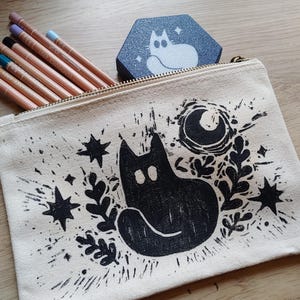 Peut inclure: Une trousse crème avec un motif de chat noir, des étoiles et une lune. La trousse est ouverte, révélant plusieurs crayons de couleur. Une petite boîte hexagonale avec un motif de chat est posée dessus.