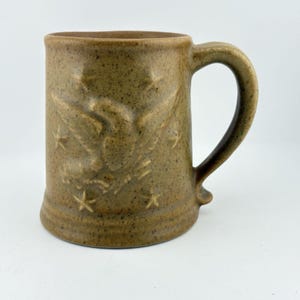 May include: A speckled brown ceramic mug featuring a raised relief design of an eagle with outstretched wings surrounded by several five-pointed stars. The mug has a cylindrical body, a sturdy handle on the right side, and a smooth, matte finish.