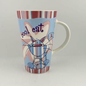 Può includere: Una tazza alta in ceramica con manico bianco. La tazza presenta uno sfondo azzurro con un'illustrazione di un gatto dei cartoni animati e le parole "cool cat". Il bordo superiore ha un motivo a righe rosse e blu.