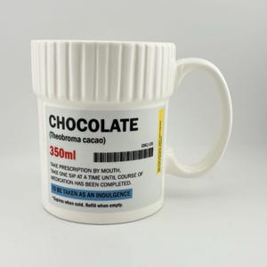 以下が含まれることがあります： 処方箋ラベルのデザインの白いセラミックマグ。 マグには、太字の「CHOCOLATE」という単語と、「（Theobroma cacao）」と「350ml」のテキストが記載されています。 マグにはハンドルとリブ付きのトップが付いています。