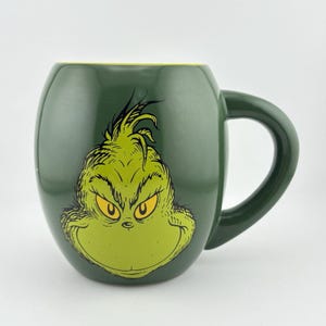 May include: Green ceramic mug featuring a cartoon image of the Grinch's face. The mug has a dark green exterior with a lighter green Grinch face and yellow eyes. The interior rim is yellow, and the handle is dark green.