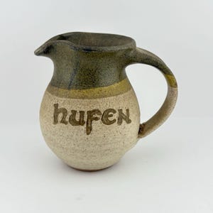 Peut inclure: Pichet en céramique avec une anse et un bec verseur. Le dessus est vert foncé, avec une bande jaune en dessous et une base beige. Le mot "hufen" est gravé sur le devant du pichet. Le pichet est sur fond blanc.