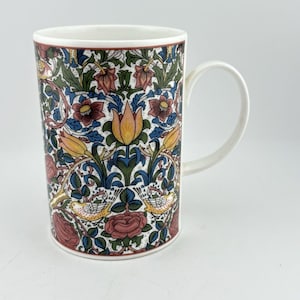 Op de afbeelding: Een keramische mok met een gedetailleerd botanisch patroon van gele tulpen, roze rozen en kleine vogels tussen groene bladeren en blauwe ranken. Het ontwerp bedekt het gehele buitenoppervlak en wordt gecompleteerd door een wit handvat en rand.