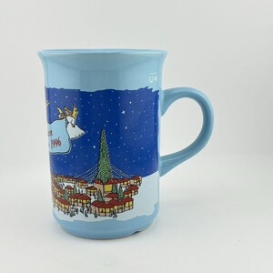 May include: A light blue ceramic mug with a handle. The mug features a winter scene with a village, a Christmas tree, and an angel carrying a banner with the year 1996. The mug has a capacity of 0.24 liters.
