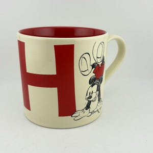 Può includere: Tazza in ceramica color crema con interno e manico rossi. La lettera "H" è visualizzata in rosso sul davanti. Un disegno in bianco e nero di Topolino è sul lato, con una camicia rossa.