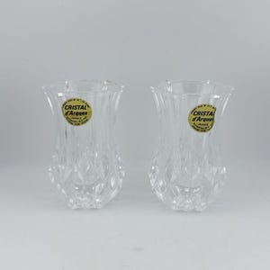 Puede incluir: Dos jarrones de cristal transparente con un patrón de corte de diamante facetado en la mitad inferior y un borde liso y acampanado. Cada jarrón presenta una pegatina redonda dorada y negra con el texto Cristal d'Arques France, indicando cristal de plomo.