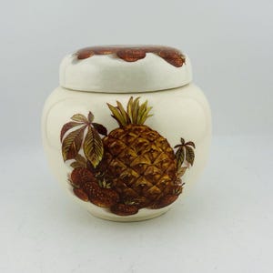 May include: A cream-colored ceramic jar with a lid, featuring a pineapple and strawberry design. The pineapple is brown and yellow, surrounded by brown leaves and strawberries. The lid has a brown, textured rim.