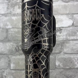 Puede incluir: Vaso t&eacute;rmico negro con dise&ntilde;o de Spiderman. El dise&ntilde;o incluye la palabra "SPIDERMAN", un patr&oacute;n de telara&ntilde;a y una imagen de Spiderman. El vaso tiene un asa y tapa negros.