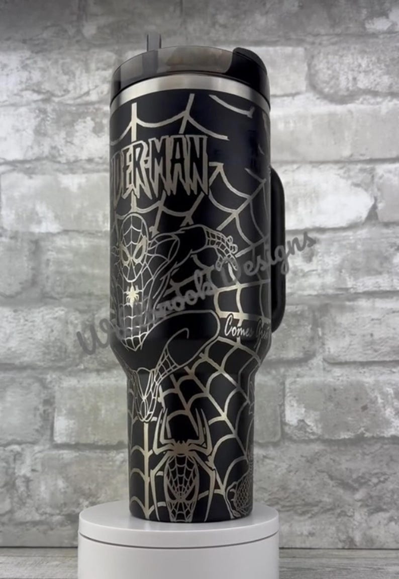 Puede incluir: Vaso t&eacute;rmico negro con dise&ntilde;o de Spiderman. El dise&ntilde;o incluye la palabra "SPIDERMAN", un patr&oacute;n de telara&ntilde;a y una imagen de Spiderman. El vaso tiene un asa y tapa negros.