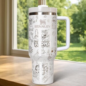 May include: White Stanley travel mug with a handle, featuring a black and white illustration of Stitch from Lilo & Stitch, with the word "Aloha". The mug has a white lid and handle. The Stanley logo is also visible.