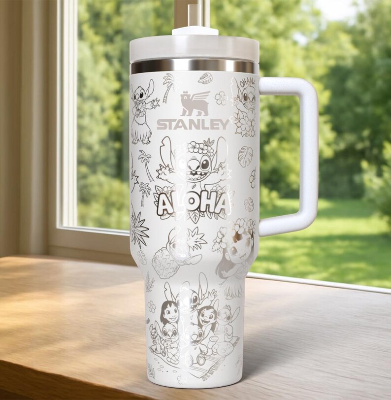 May include: White Stanley travel mug with a handle, featuring a black and white illustration of Stitch from Lilo & Stitch, with the word "Aloha". The mug has a white lid and handle. The Stanley logo is also visible.