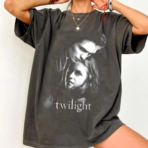 May include: Dark gray t-shirt featuring a black and white image of two people with the word "twilight" in white text. The shirt has short sleeves and a relaxed fit.