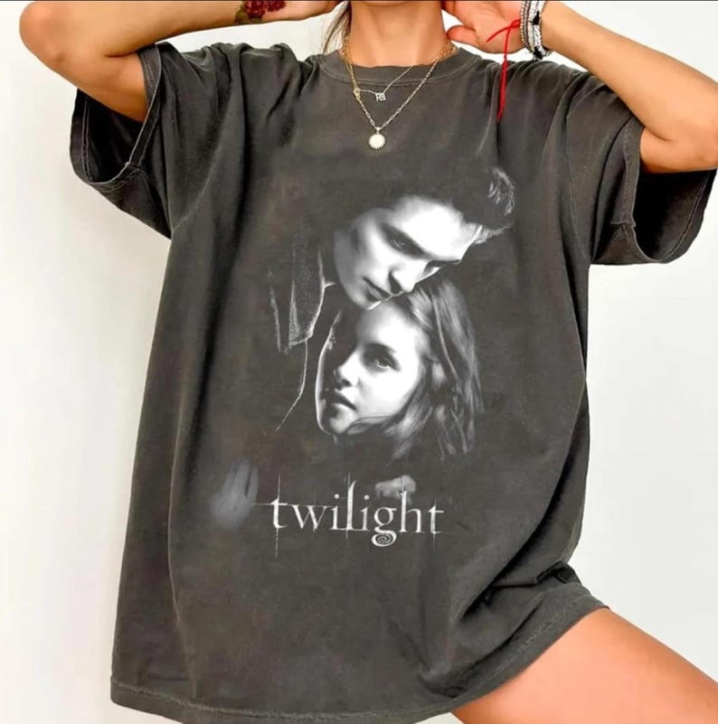 May include: Dark gray t-shirt featuring a black and white image of two people with the word "twilight" in white text. The shirt has short sleeves and a relaxed fit.