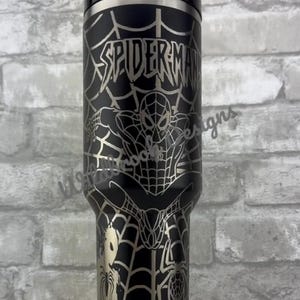 Puede incluir: Vaso de acero inoxidable negro con un dise&ntilde;o de Spider-Man. El vaso tiene una tapa y una pajita plateadas. El dise&ntilde;o incluye la palabra "SPIDERMAN" y una imagen de Spider-Man en una telara&ntilde;a. El vaso est&aacute; sobre un soporte blanco.