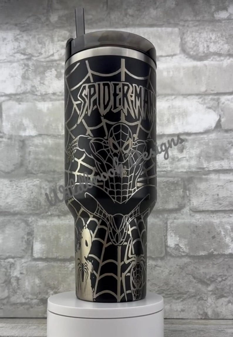 Puede incluir: Vaso de acero inoxidable negro con un dise&ntilde;o de Spider-Man. El vaso tiene una tapa y una pajita plateadas. El dise&ntilde;o incluye la palabra "SPIDERMAN" y una imagen de Spider-Man en una telara&ntilde;a. El vaso est&aacute; sobre un soporte blanco.