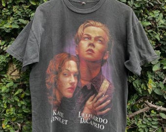 May include: Dark gray t-shirt featuring a graphic of Kate Winslet and Leonardo DiCaprio. The names of the actors are printed in white. The shirt is hanging on a wooden hanger against a green leafy background.
