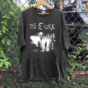 May include: Dark gray t-shirt featuring the band name "THE CURE" in white, above a black and white graphic of a street scene with a figure under a streetlight. The shirt is hanging on a wooden hanger.