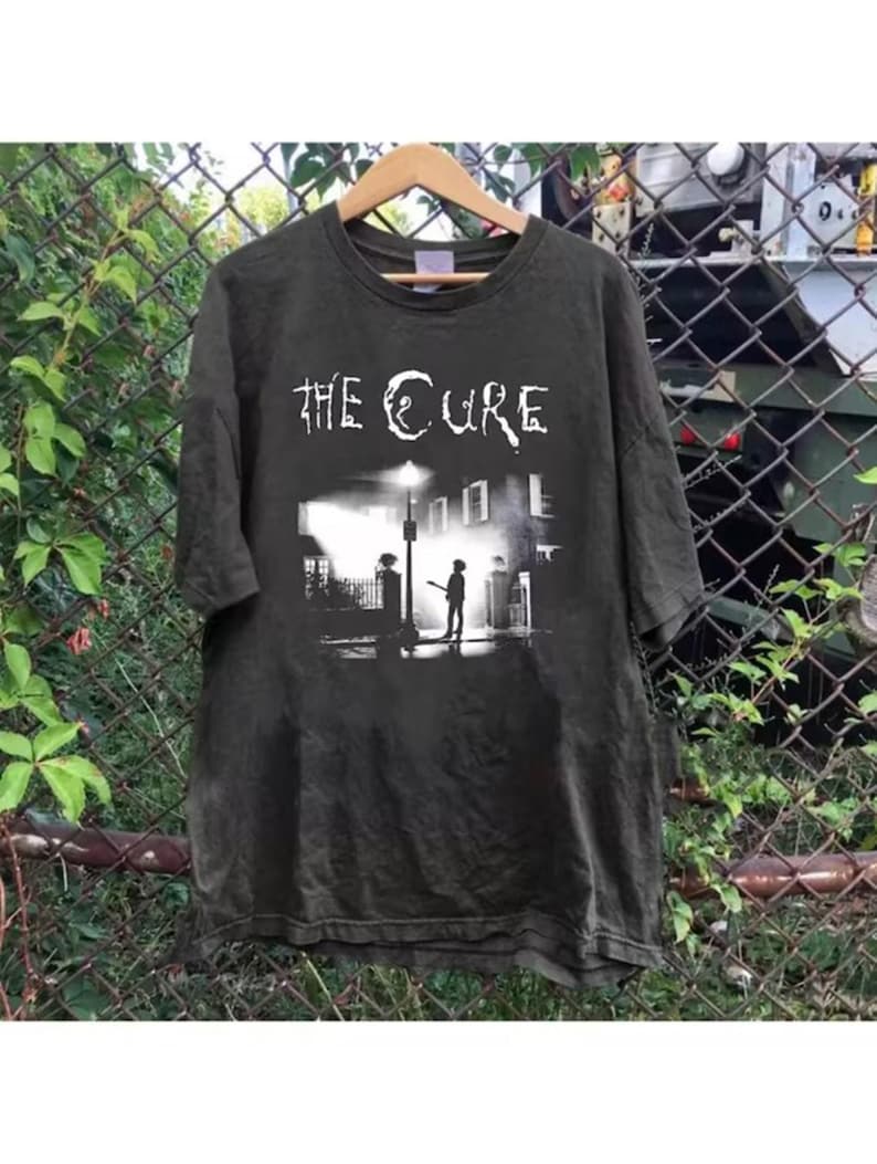May include: Dark gray t-shirt featuring the band name "THE CURE" in white, above a black and white graphic of a street scene with a figure under a streetlight. The shirt is hanging on a wooden hanger.