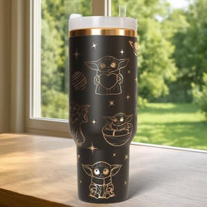 May include: Black tumbler with gold accents and a white lid. The tumbler features gold illustrations of a character holding a heart, in a pod, and other designs. The tumbler has a gold band near the top and a straw.