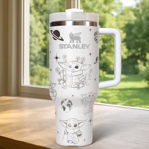 May include: White insulated tumbler with a handle and lid. The tumbler features a design of Baby Yoda, planets, stars, and flowers. The Stanley logo is printed on the front. The tumbler is sitting on a wooden surface.