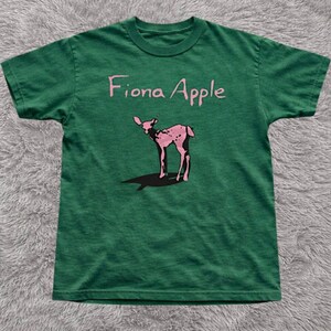 May include: Green t-shirt featuring the text "Fiona Apple" above a pink deer graphic. The deer has a spotted pattern and a long shadow. The shirt is made of a soft material.