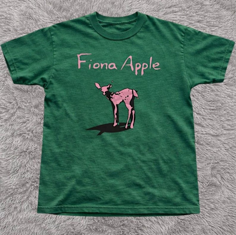 May include: Green t-shirt featuring the text "Fiona Apple" above a pink deer graphic. The deer has a spotted pattern and a long shadow. The shirt is made of a soft material.