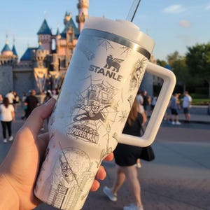 Puede incluir: Un vaso Stanley blanco con un dise&ntilde;o de Spider-Man, un asa y una pajita. El vaso tiene un borde plateado y una tapa blanca. Los gr&aacute;ficos de Spider-Man incluyen telara&ntilde;as, paisajes urbanos y el logotipo de Spider-Man.