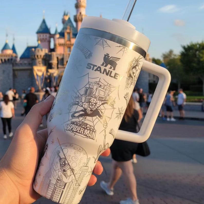 Puede incluir: Un vaso Stanley blanco con un dise&ntilde;o de Spider-Man, un asa y una pajita. El vaso tiene un borde plateado y una tapa blanca. Los gr&aacute;ficos de Spider-Man incluyen telara&ntilde;as, paisajes urbanos y el logotipo de Spider-Man.