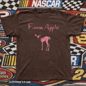 May include: Brown t-shirt featuring the text "Fiona Apple" in pink script above a pink deer graphic. The shirt is a solid brown color and has short sleeves.