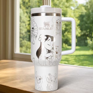 May include: White Stanley tumbler with a handle, featuring a design of Lilo & Stitch characters. The tumbler has a white lid and handle, with the Stanley logo. The design includes illustrations of Lilo, Stitch, and other characters from the movie.